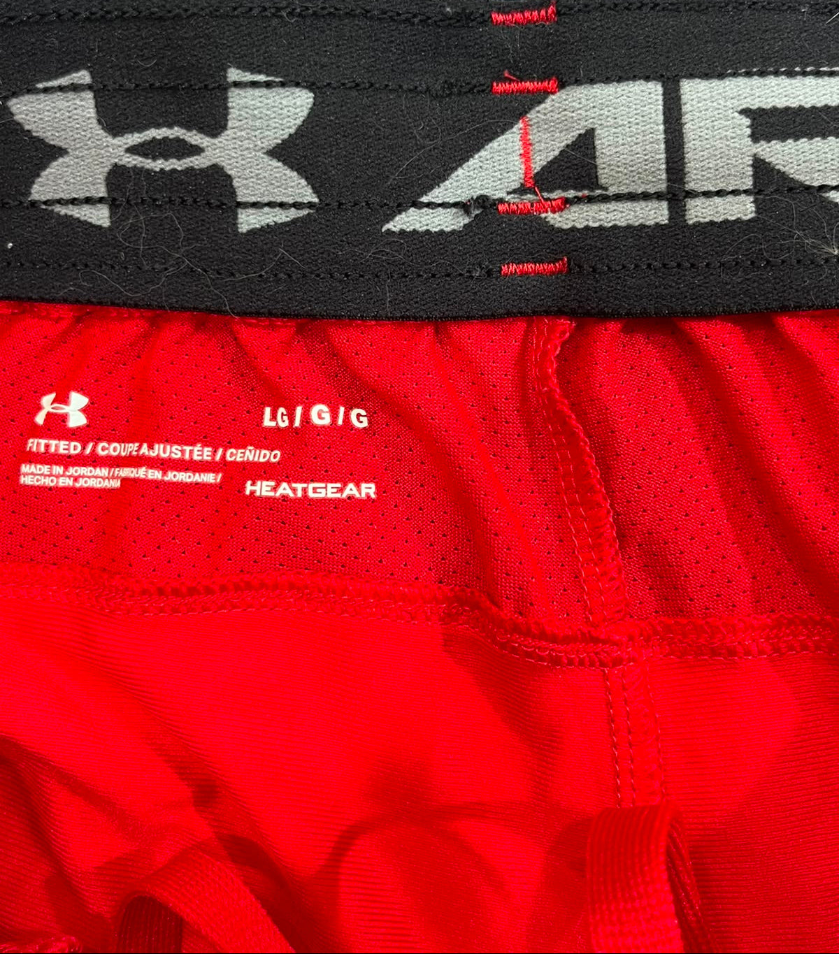 Close-up of red athletic shorts showing the inner label with Under Armour branding and size information, alongside a black waistband with a large gray Under Armour logo.