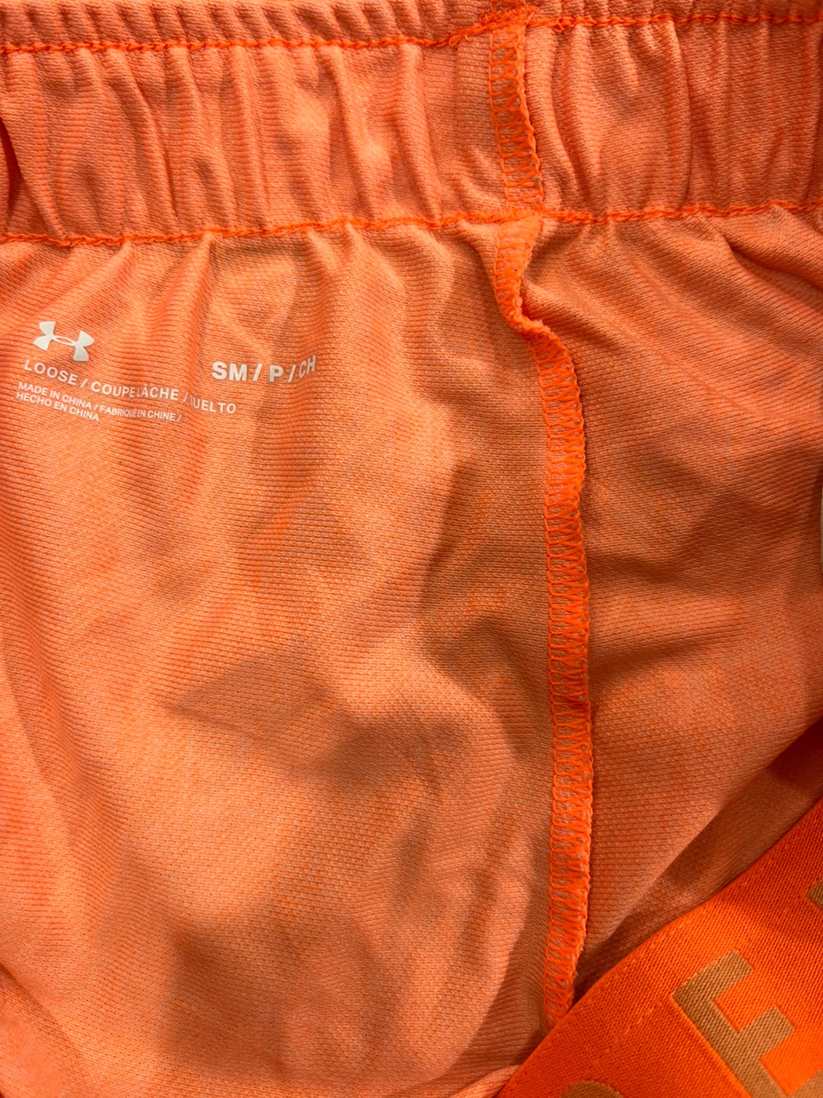 Close-up of the inside waistband of orange athletic shorts, showing the elasticized orange fabric, white Under Armour logo and size text, and a matching orange drawstring.