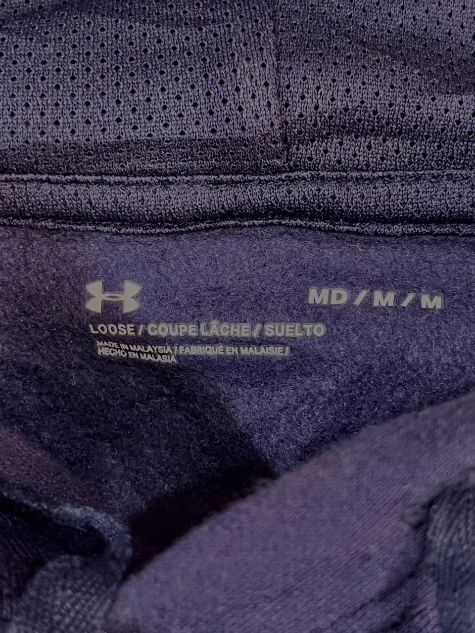 Close-up of the inner neck label of a dark purple athletic top, showing the Under Armour logo and size information 'MD / M / M'.