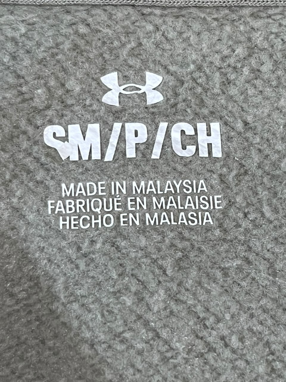 Close-up of a gray textured garment interior label with the Under Armour logo and white text reading 'SM/P/CH' and 'MADE IN MALAYSIA'.