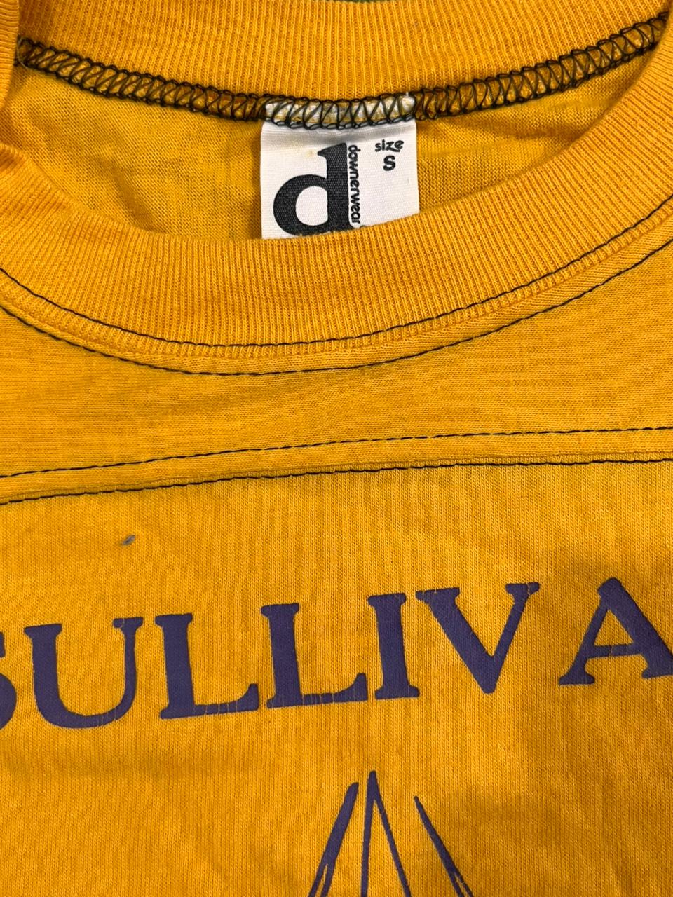 Close-up of a mustard yellow crewneck t-shirt with dark blue text reading 'SULLIVA' and a white neck label showing the brand 'd'comfyt' and size 'S'.