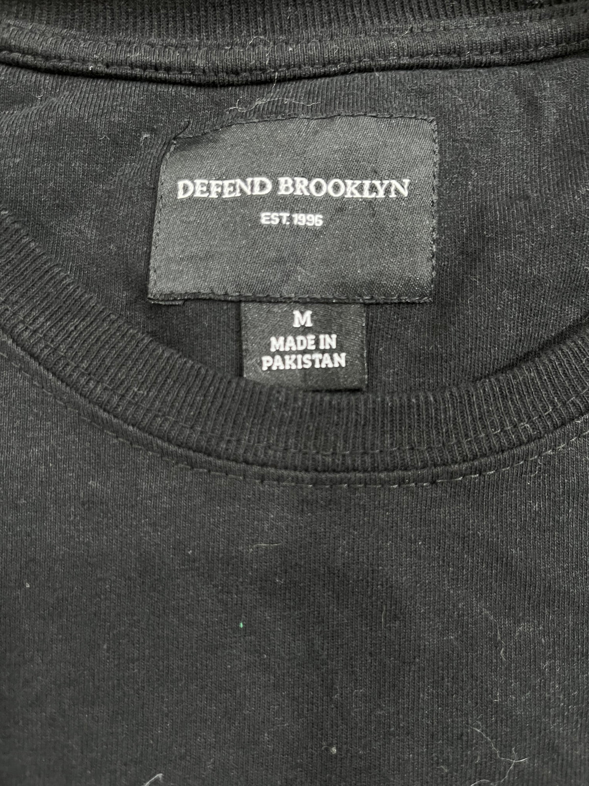 Close-up of the inside neck of a black crewneck sweatshirt, showing the 'DEFEND BROOKLYN' brand label and a 'MADE IN PAKISTAN' size tag.