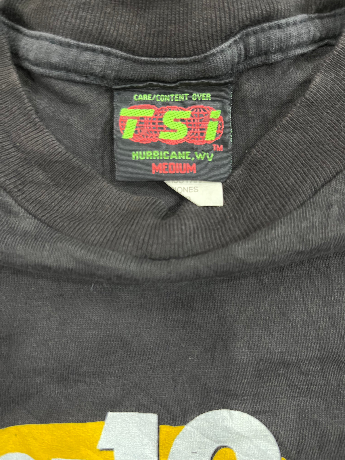Close-up of the inside neck label of a dark gray T-shirt, showing the brand 'TSl' with 'HURRICANE, wv MEDIUM' printed on the black tag.