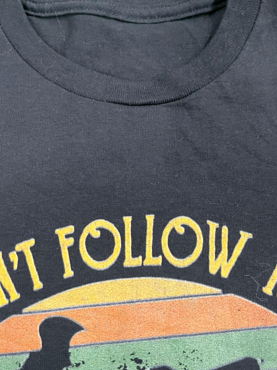 Close-up of a dark charcoal gray t-shirt featuring a vintage-style graphic with yellow text reading 'I DON'T FOLLOW' above a retro sunset design in yellow, orange, green, and black.
