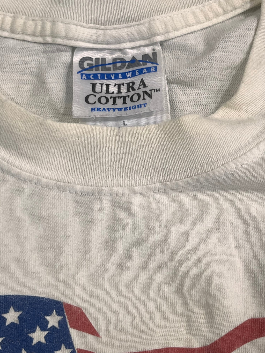Close-up of a white Gildan Activewear Ultra Cotton Heavyweight t-shirt showing the inner neck label and part of a red, white, and blue American flag graphic on the right side.
