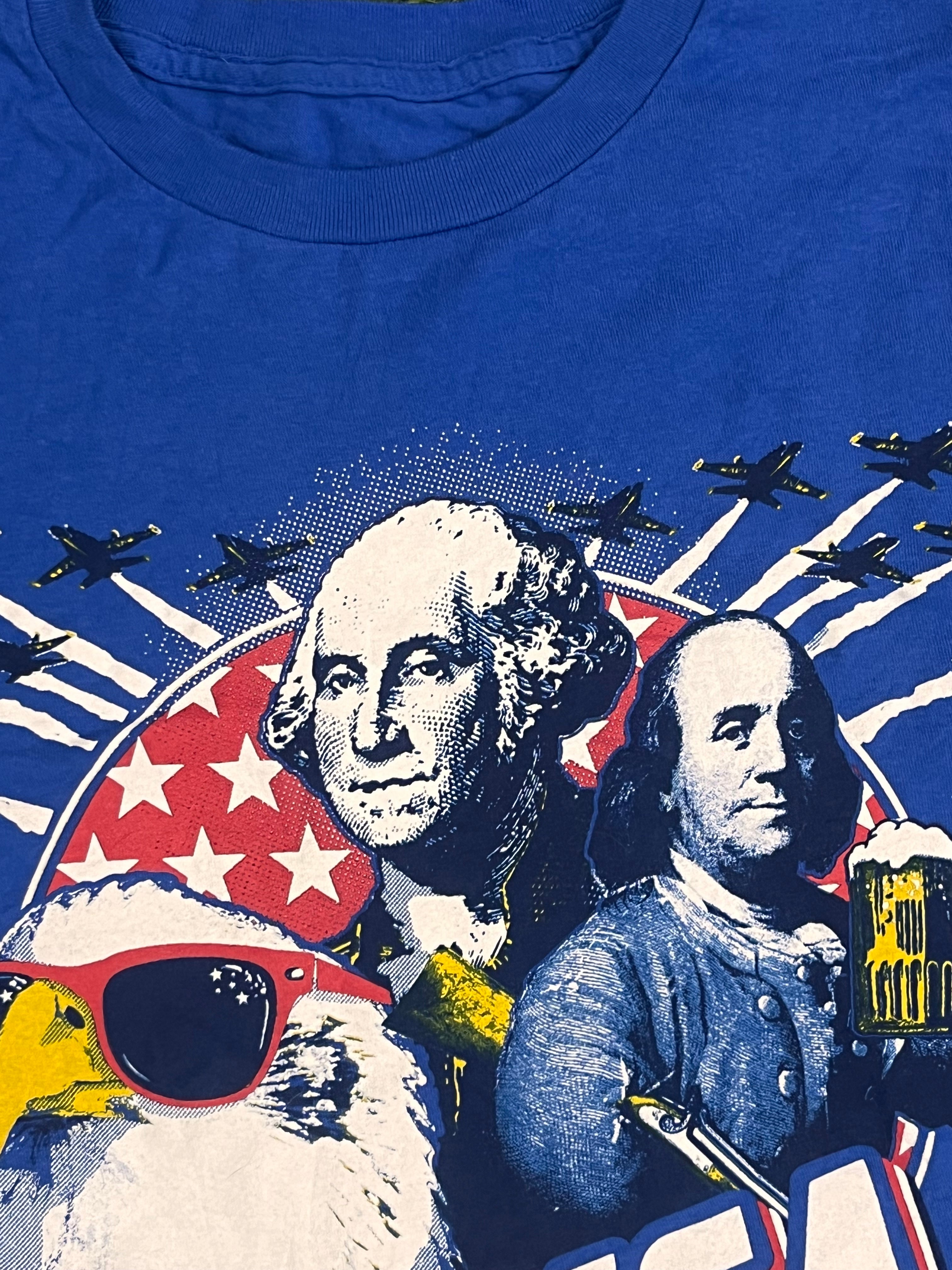 Close-up of a blue graphic t-shirt featuring a stylized print of George Washington, a dog in a spacesuit with red sunglasses, jets, and an American flag motif.