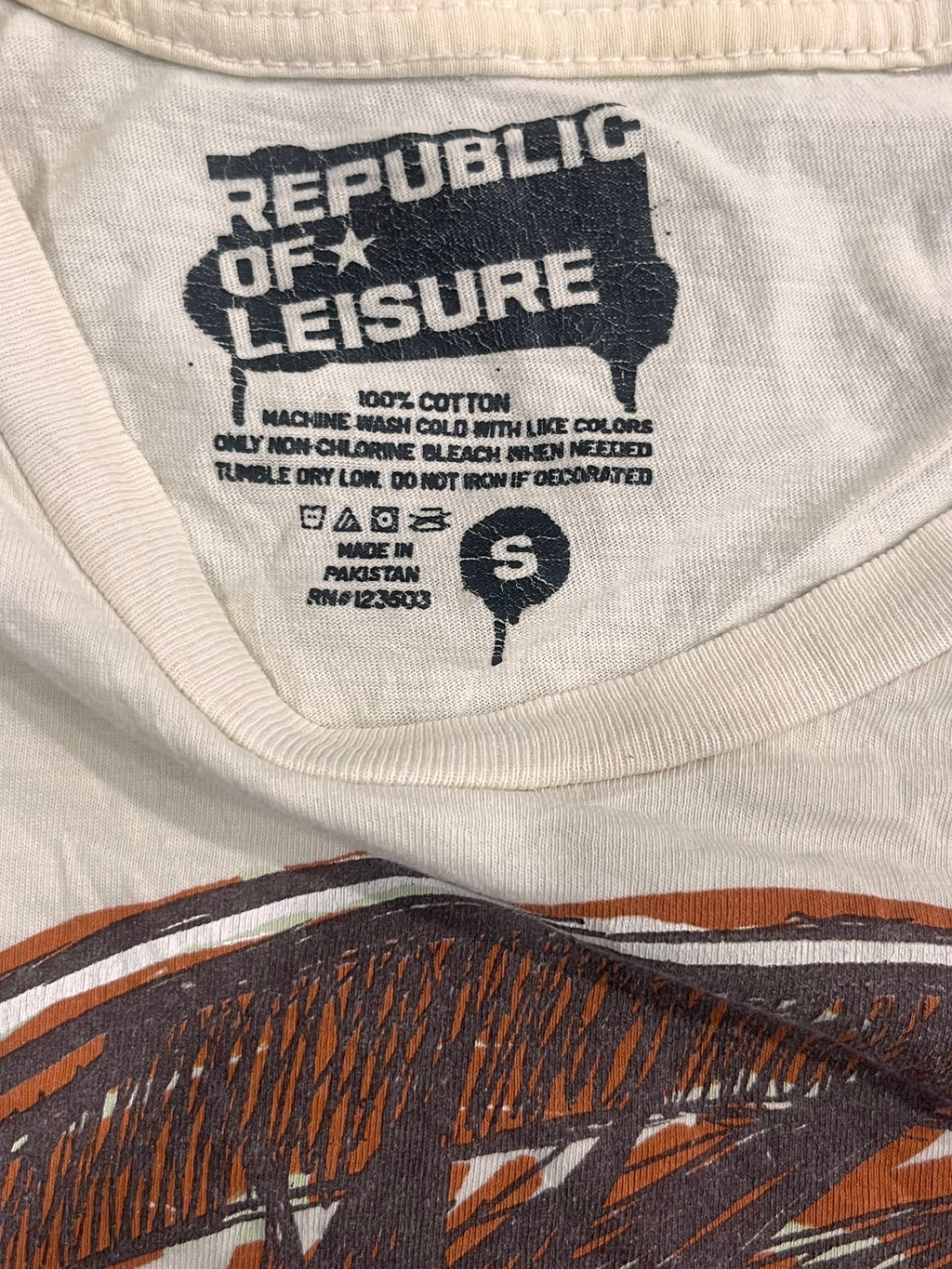 Close-up of the inside collar of a white cotton t-shirt by Republic of Leisure, showing the black printed brand logo, care instructions, and a size label 'S'. The bottom of the shirt features a brown and black graphic print.