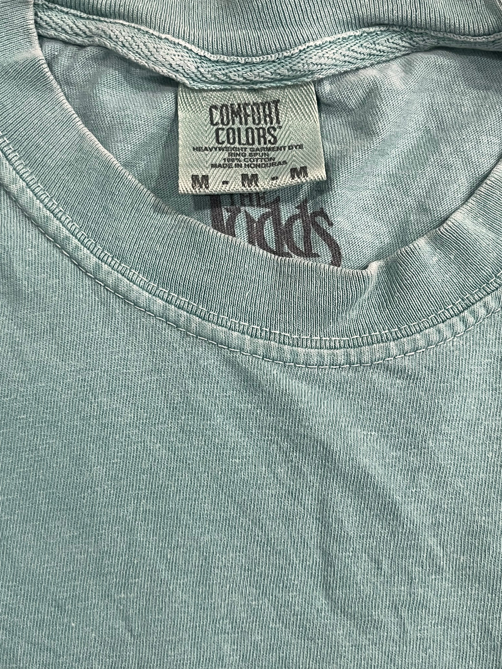 Close-up of a light sage green t-shirt showing the inner neck label which reads 'COMFORT COLORS' and 'MY / ME'.