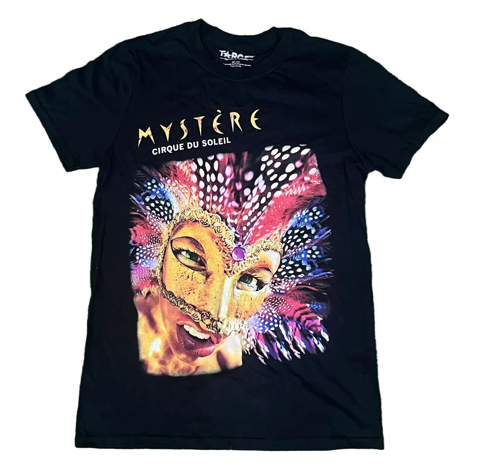 A black short-sleeve graphic T-shirt . The shirt features the words 'MYSTÈRE CIRQUE DU SOLEIL' in gold and white above a large colorful carnival mask illustration with pink, purple, blue, and gold tones.