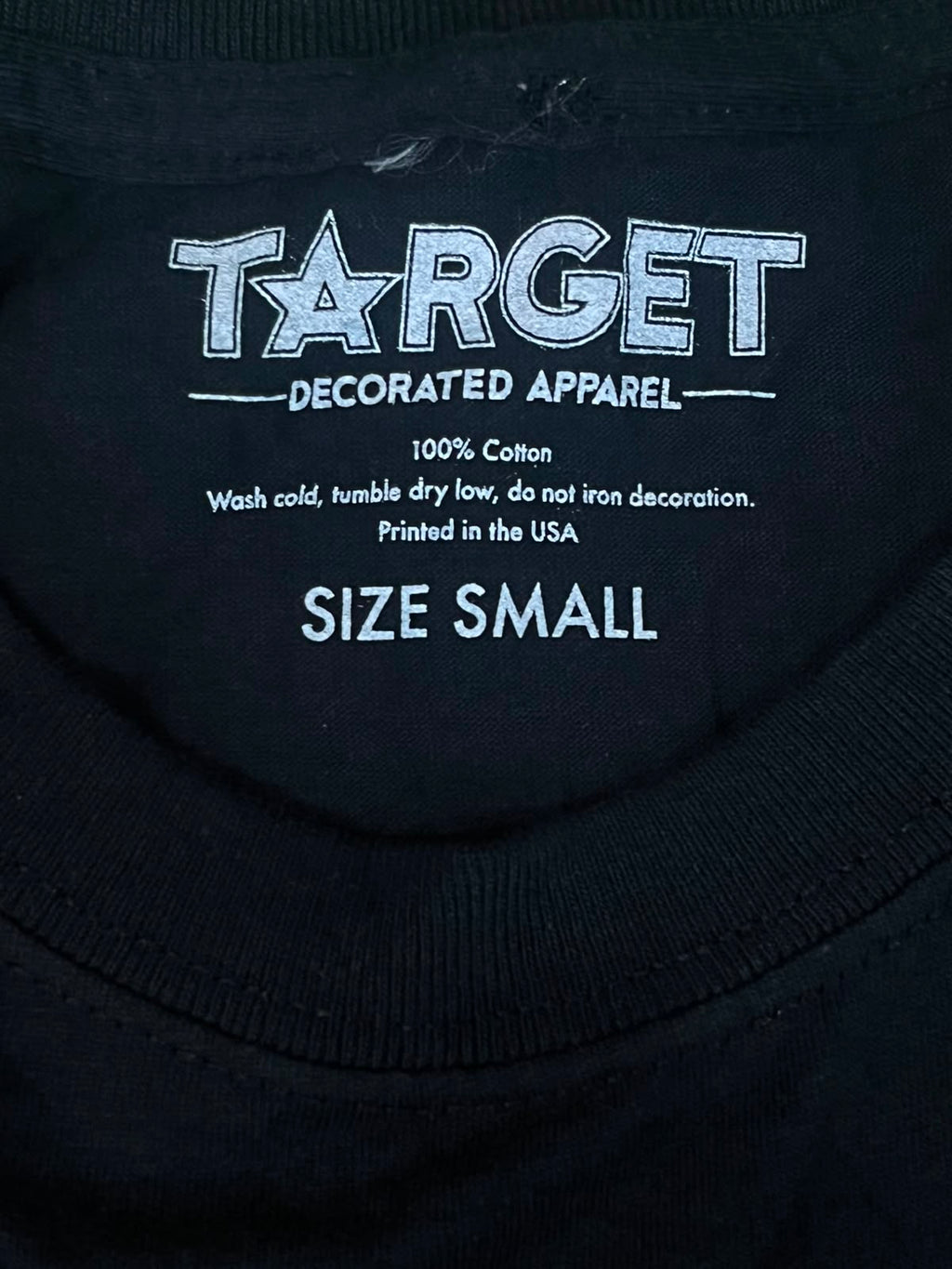 Close-up of the inside collar of a black T-shirt showing printed white text that reads TARGET DECORATED APPAREL, 100% Cotton, Wash cold, tumble dry low, do not iron decoration, Printed in the USA, and SIZE SMALL.