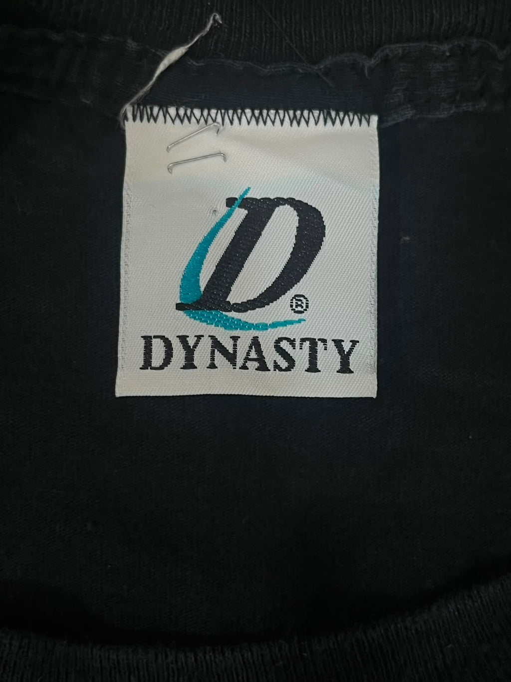 Close-up of a black garment's inner neck label featuring a white patch with a stylized black 'D' logo and the brand name 'DYNASTY' in black text.