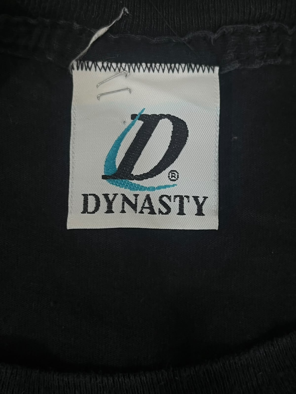 Close-up of a black garment's inner neck label featuring a white patch with a stylized black 'D' logo and the brand name 'DYNASTY' in black text.