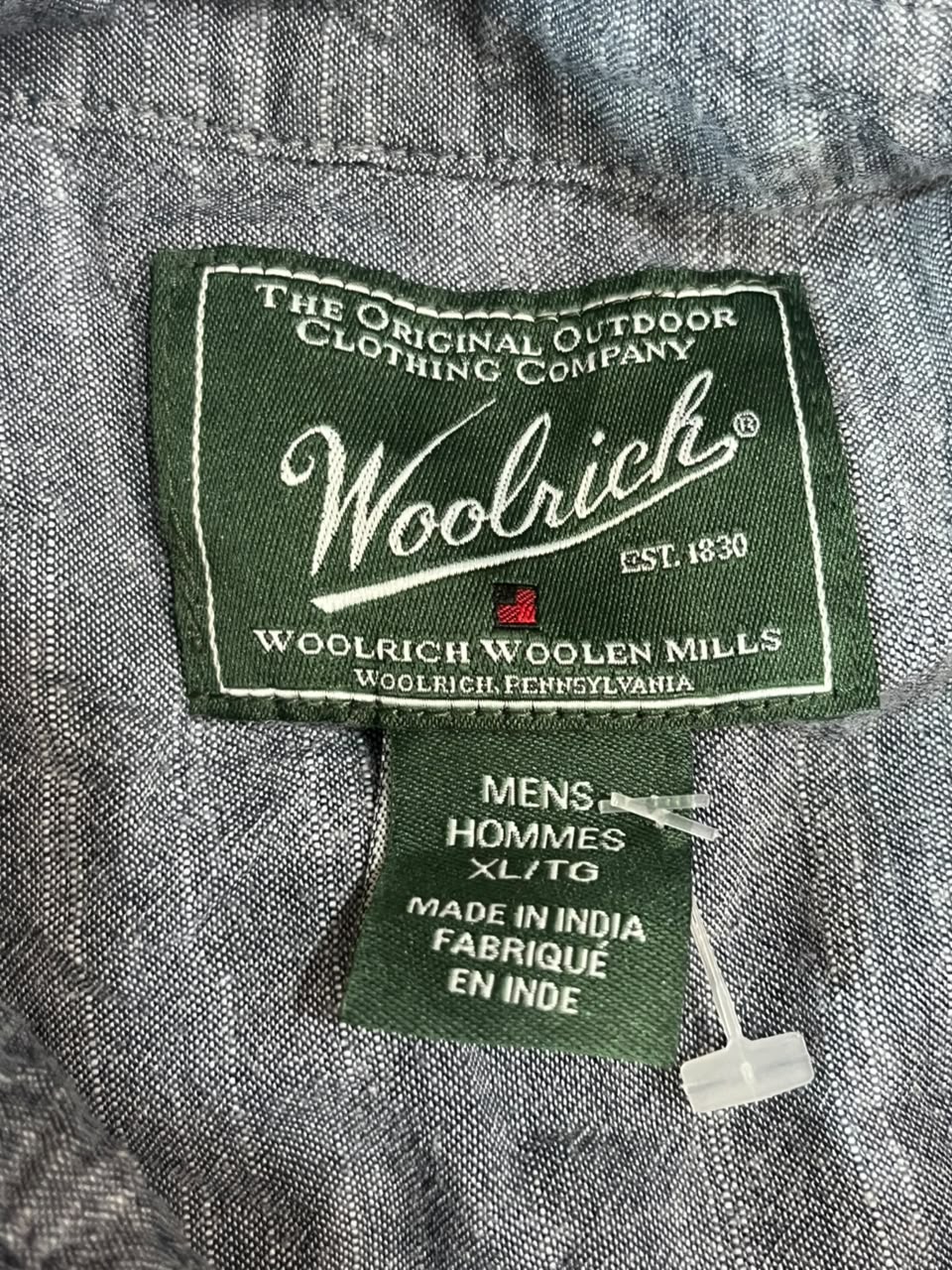 Close-up of a gray woolen clothing label reading Woolrich, with a smaller green tag below that says Mens, Hommes, XL/TO, Made in India, Fabrique En Inde.