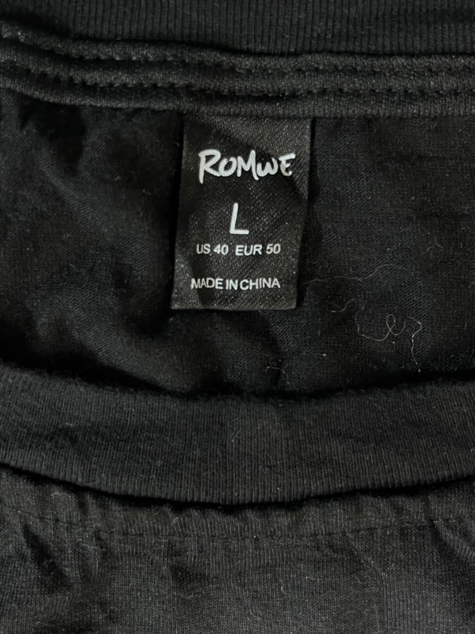 Close-up of a black garment's inner neck label reading ROMWE, size L, with US 40 / EUR 50 and MADE IN CHINA printed in white text.