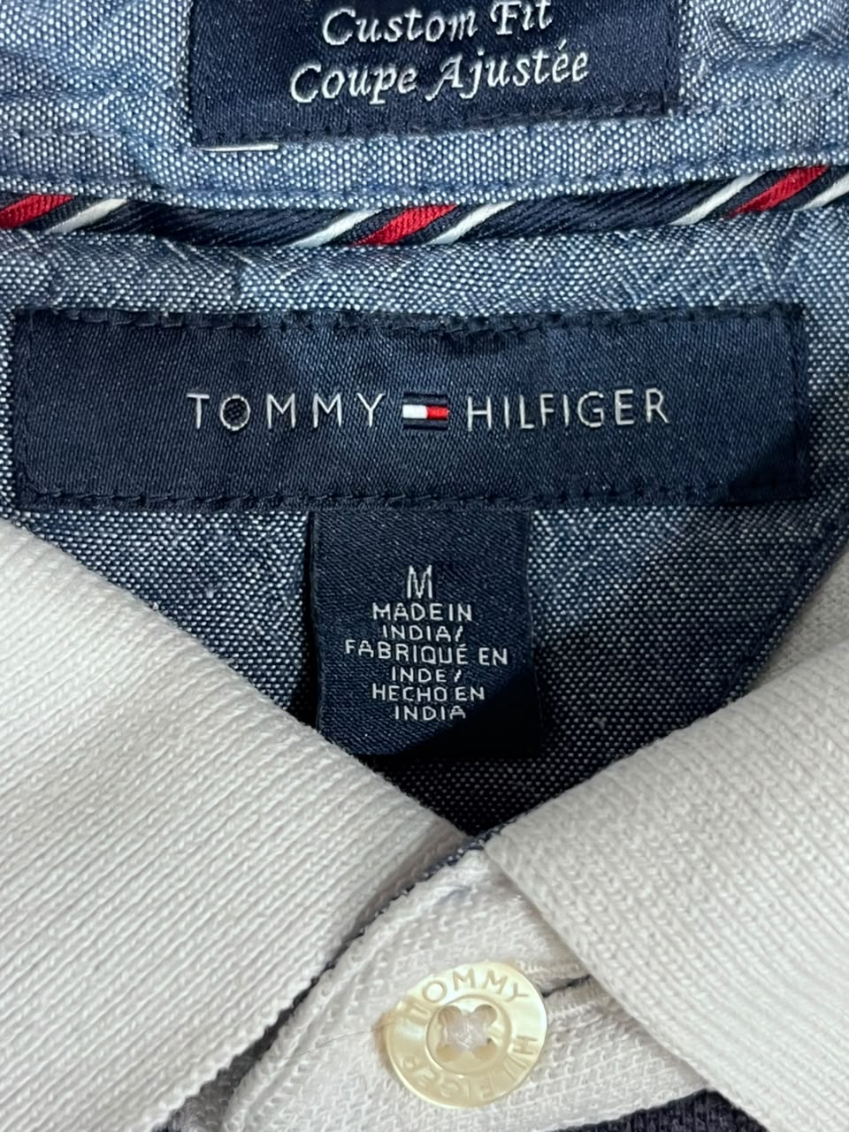 Close-up of a Tommy Hilfiger custom fit collared shirt showing the navy blue inner neck label with the brand name, a size tag reading M, and part of the light-colored knit collar.