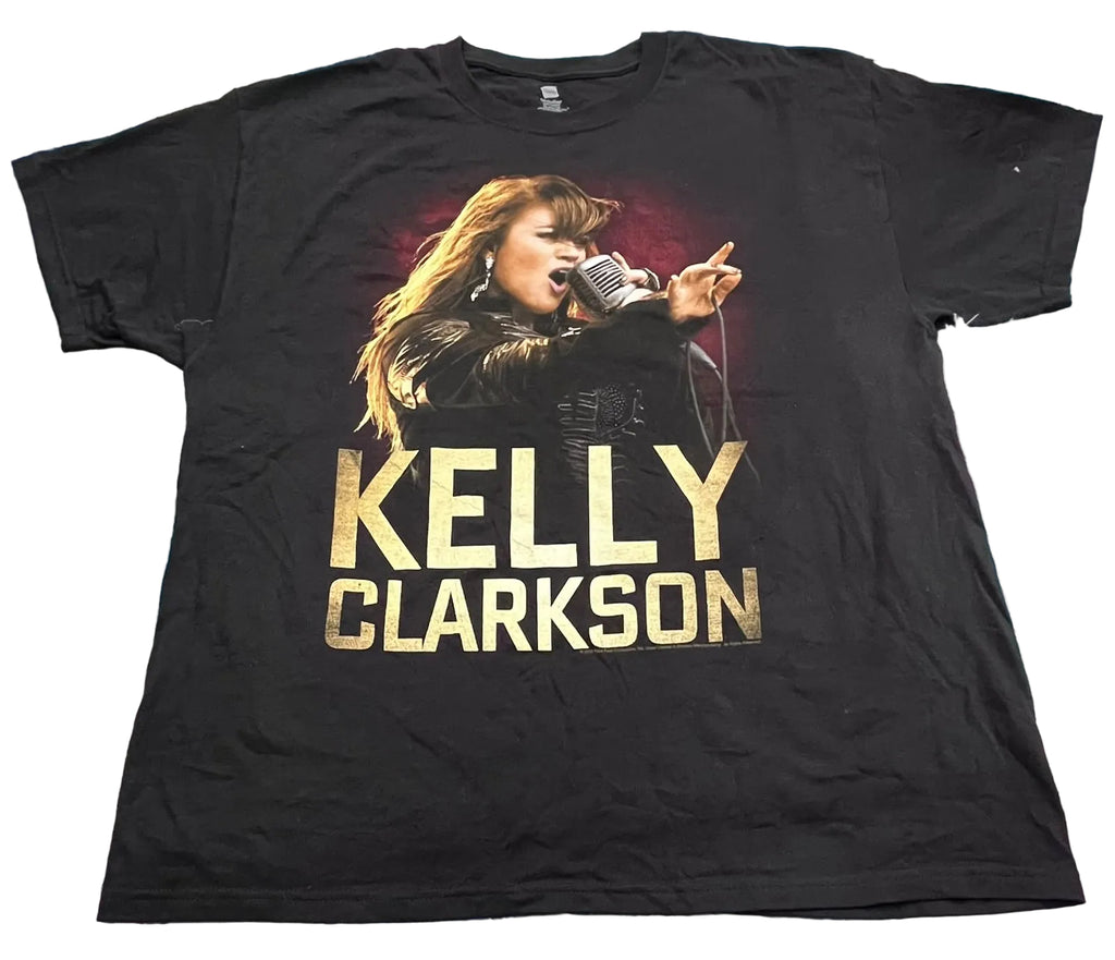 A black short-sleeve graphic T-shirt. The shirt features a large front print of Kelly Clarkson singing into a microphone, with 'KELLY CLARKSON' printed below in large gold letters.