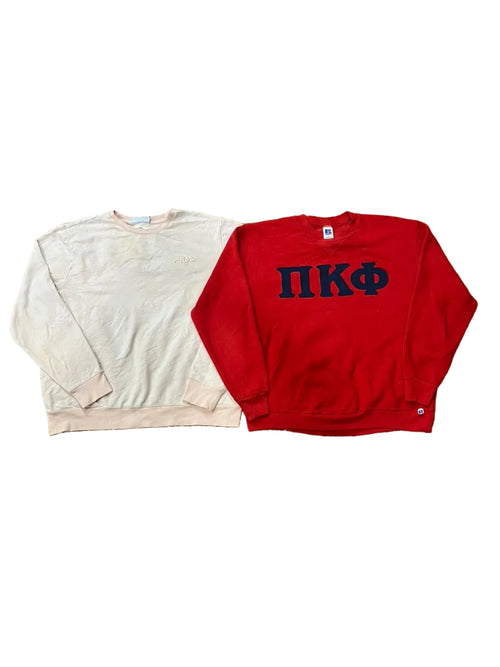Two sweatshirts a light cream sweatshirt on the left and a bright red sweatshirt on the right with dark navy Greek letters 'ΠΚΦ' across the chest. Bare feet are visible at the bottom, and other clothes are scattered around the scene.