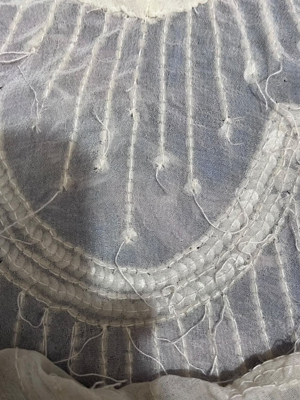 A close-up of a light gray or silver-gray sheer mesh fabric with white embroidery, featuring vertical stitched lines and a decorative scalloped border.