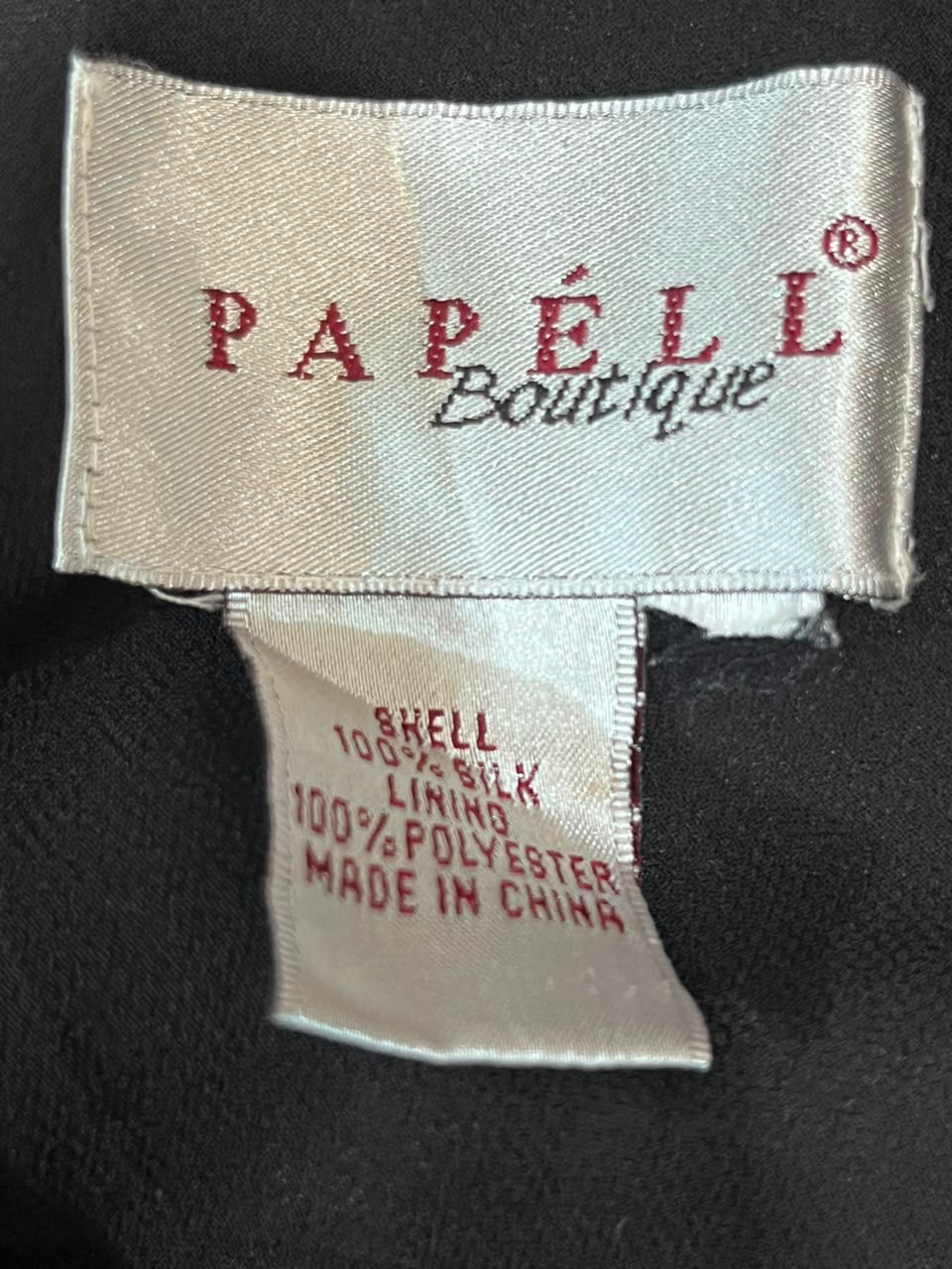 Close-up of a garment label showing the brand 'Papél Boutique' in red script on a white fabric tag, next to a size and material tag on a black textured background.