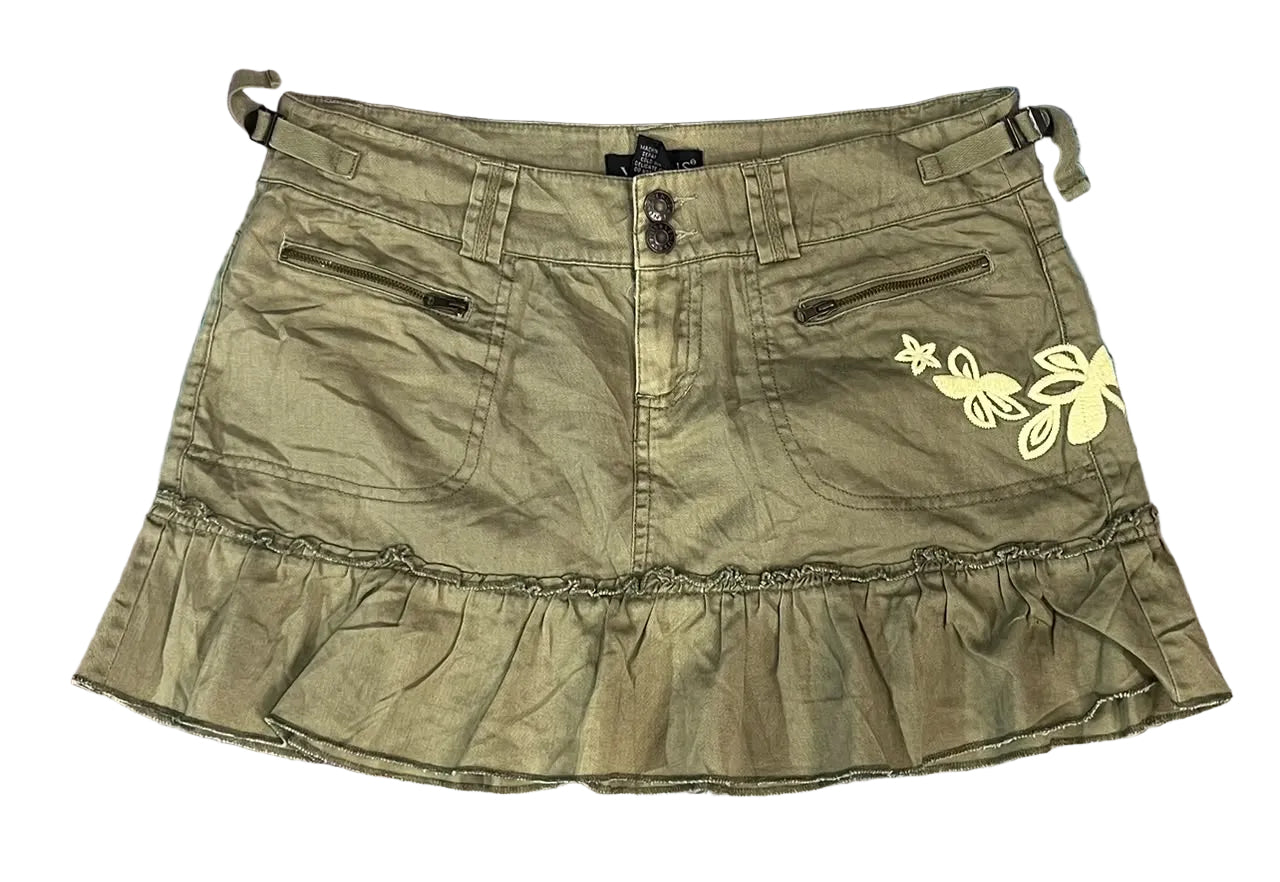 An olive green denim mini skirt with a ruffled hem, front button closure, zippered pockets, and a yellow embroidered butterfly design on the left side,
