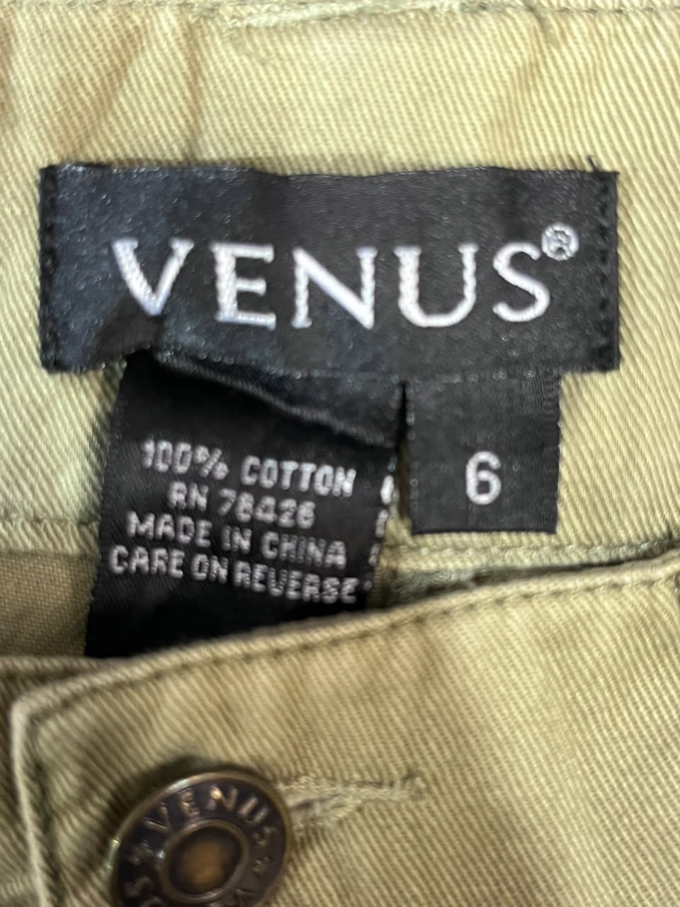 Close-up of a garment label reading VENUS, with additional fabric tags showing 100% cotton, size 6, and made in China, on a beige woven textile.