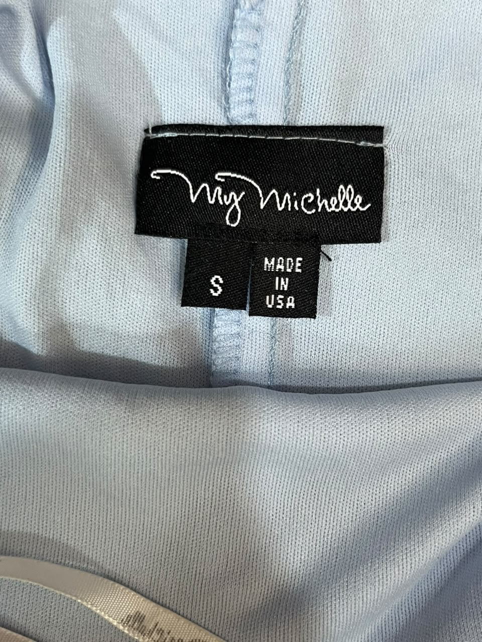 Close-up of a light blue garment's inner neck label, showing a black brand tag reading 'My Michelle' and a size tag marked 'S'.