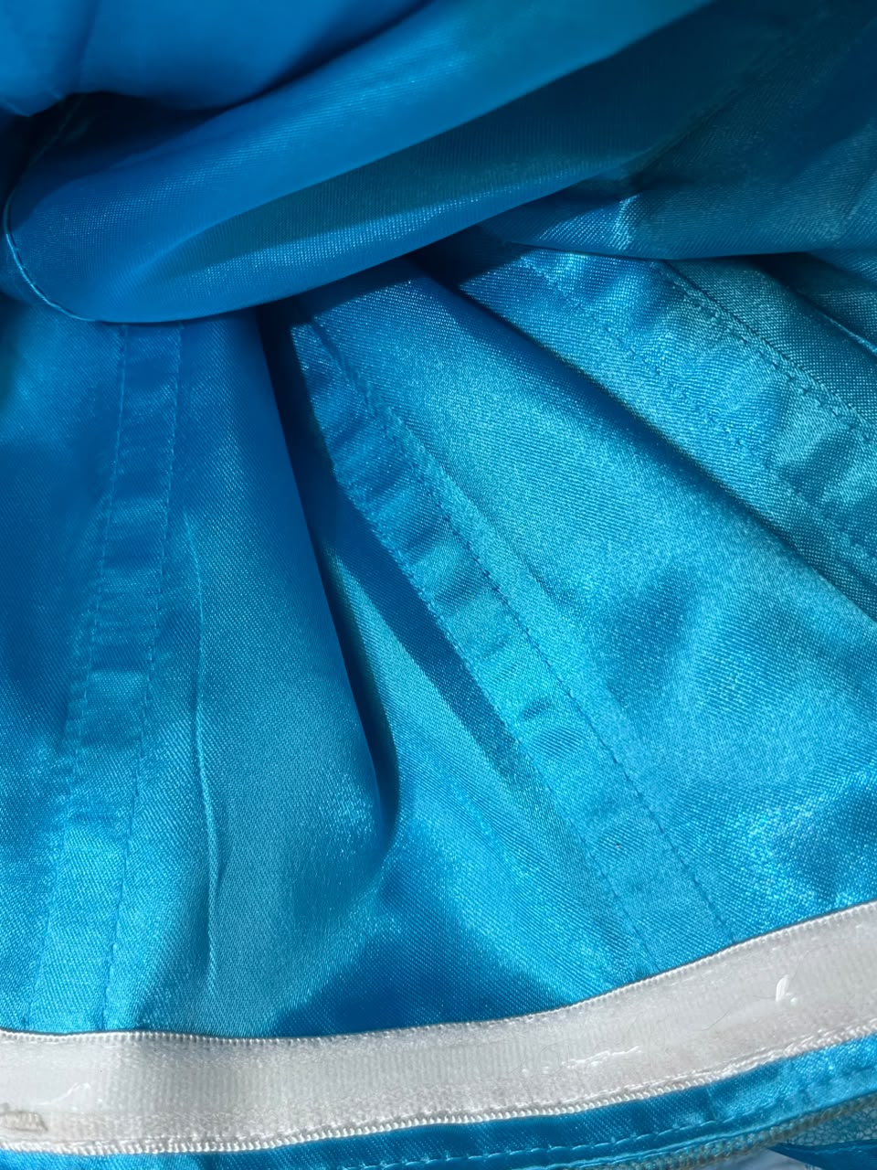 A close-up of a bright blue satin-like fabric with visible pleats and a light-colored trim along the bottom edge.