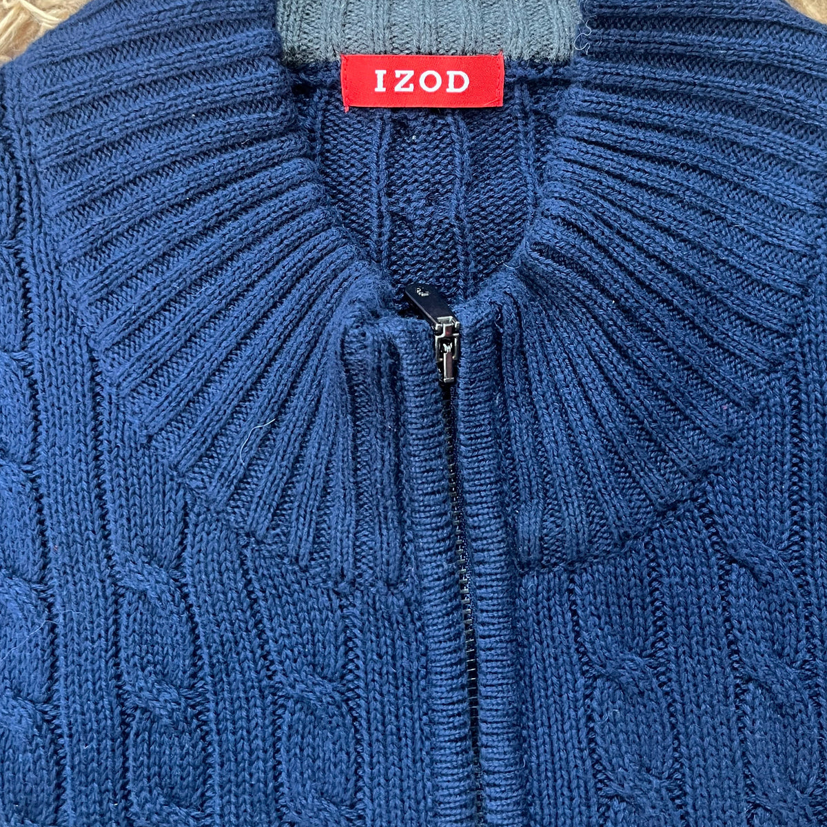 A close-up of a blue cable-knit zip-up sweater laid flat on a beige shag rug, with a red neck label reading IZOD.