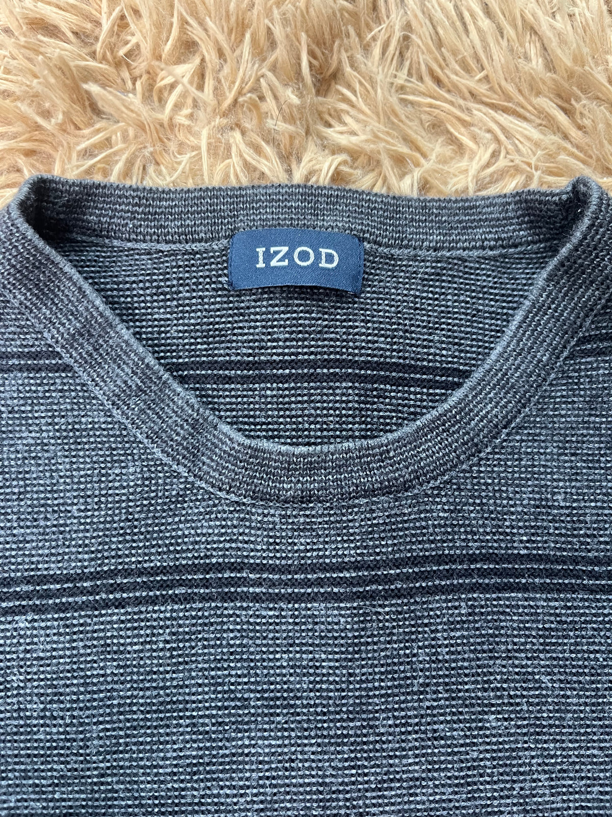 Close-up of a dark heather gray knit sweater with thin navy horizontal stripes, featuring a blue neck label with the brand name 'IZOD' in white, laid flat on a beige shag rug.