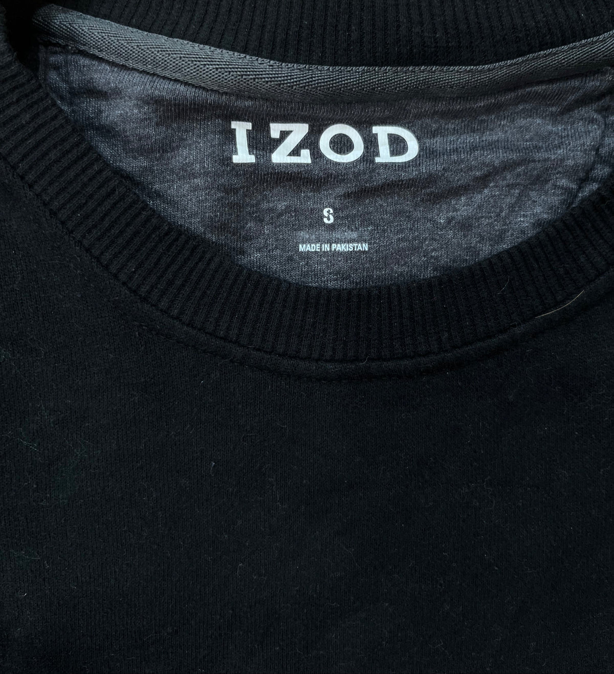 Close-up of a black crewneck sweater with the brand name 'IZOD' printed in white on the inner neck label, set against a textured beige rug background.