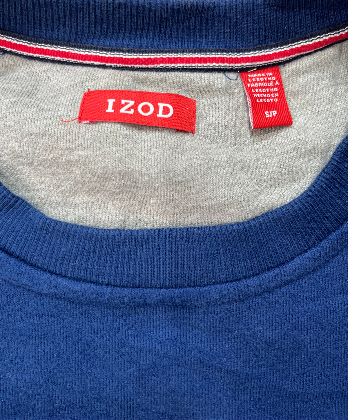 Close-up of the inner neck area of a blue knit sweater, showing a red Izod brand label and a smaller red care tag on a light gray inner collar, with a textured tan surface at the top.
