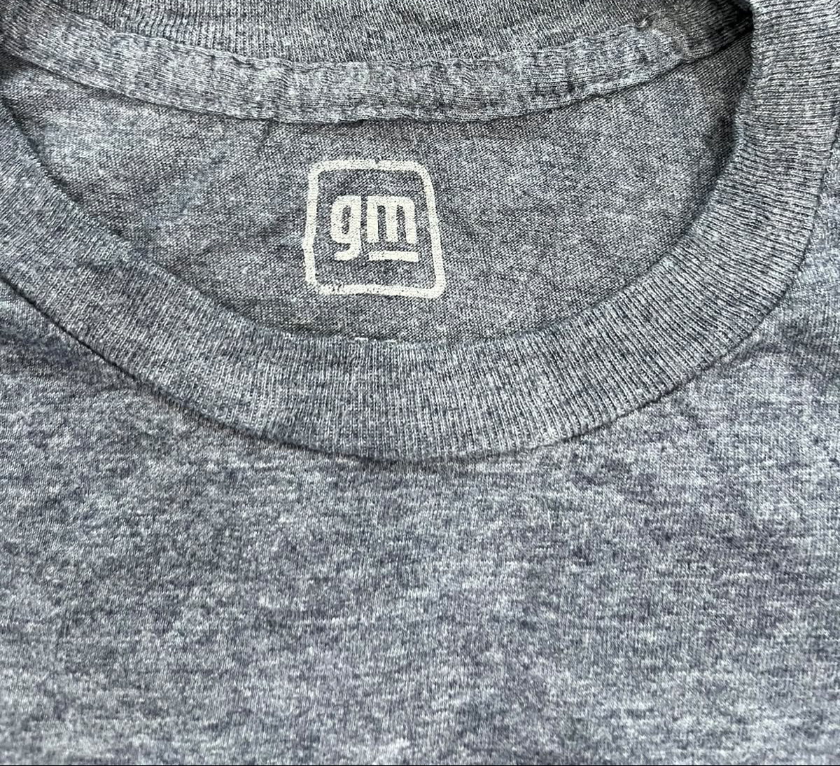 Close-up of a heather gray crewneck t-shirt laid flat on a textured olive green rug, featuring a white embroidered 'gm' logo inside the collar.